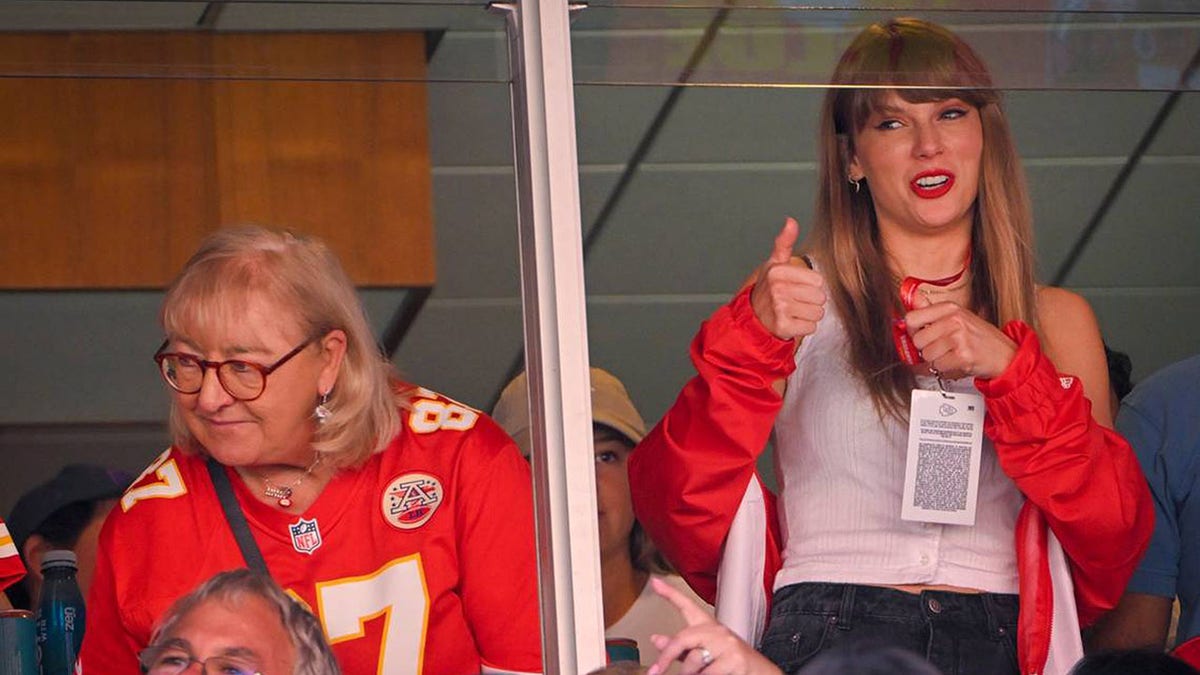 Taylor Swift in a white tank top and red jacket gives a thumbs up while standing in a suite next to Donna Kelce wearing a Travis Kelce Chiefs jersey.