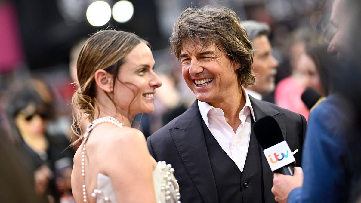 Tom Cruise in a black suit looks longingly at Rebecca Ferguson on the carpet as they're interviewed