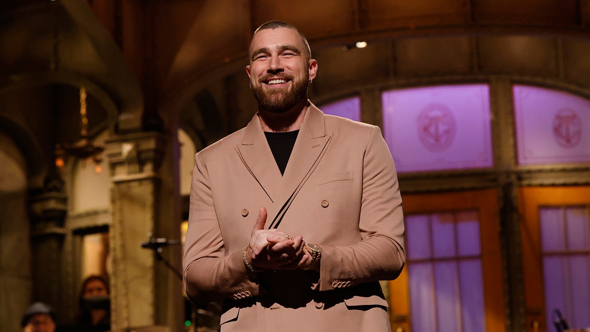 Travis Kelce holds his hands together in a tan suit as he stands on stage to host SNL