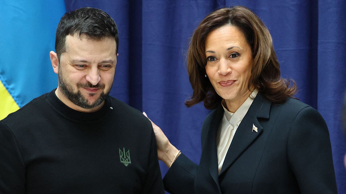 Ukrainian flag behind Ukrainian President Volodymyr Zelenskyy looking down, giving a soft closed mouth smile, U.S. Vice President Kamala Harris standing next to him, hand on shoulder, looking up giving a soft closed mouth smile