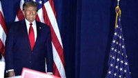Donald Trump smiling, standing in front of and next to American flags