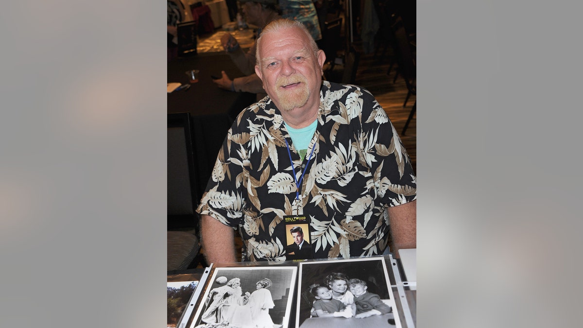 Johnny Whitaker sentado em frente a várias fotografias em preto e branco