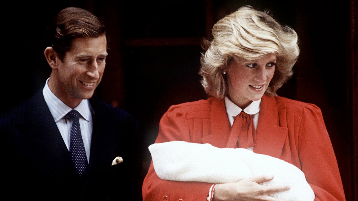 King Charles in a black suit stands next to Princess Diana in a orange/red suit holding newborn Prince Harry