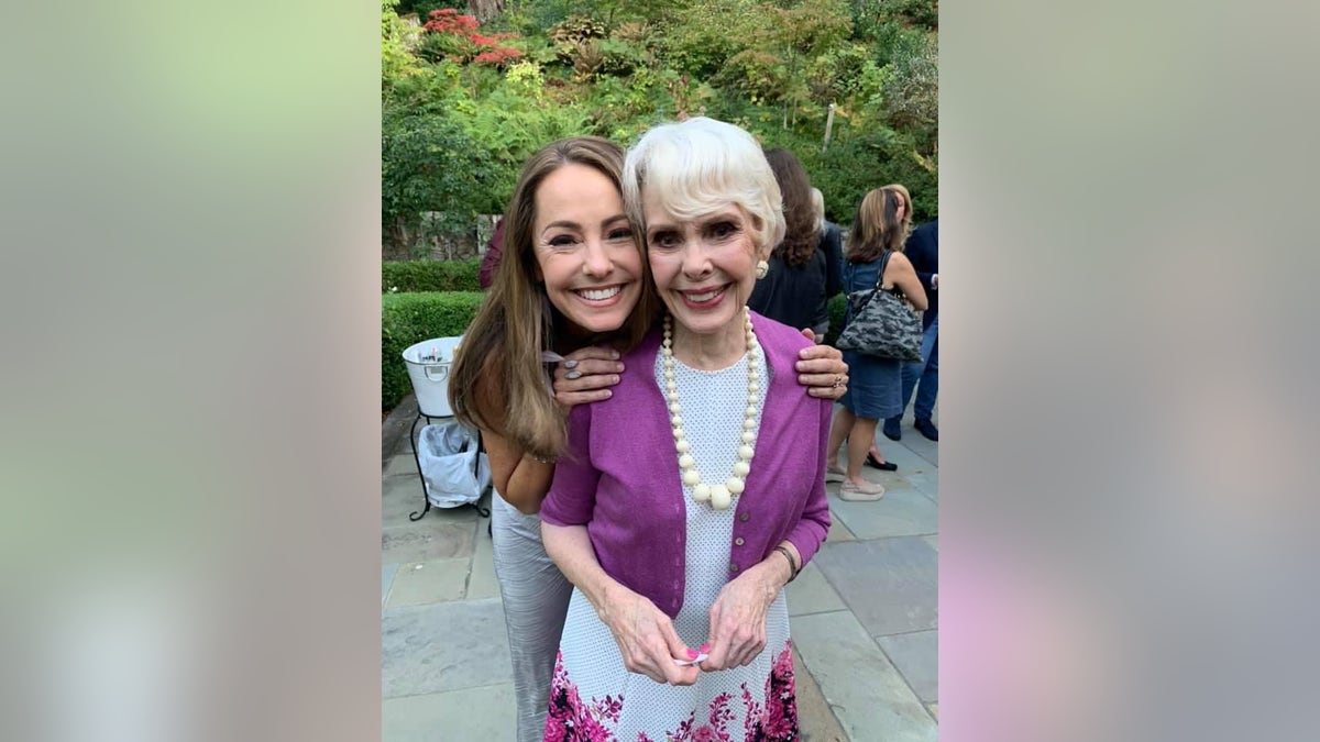 Barbara Rush wearing a white dress and purple cardigan being hugged by her daughter.