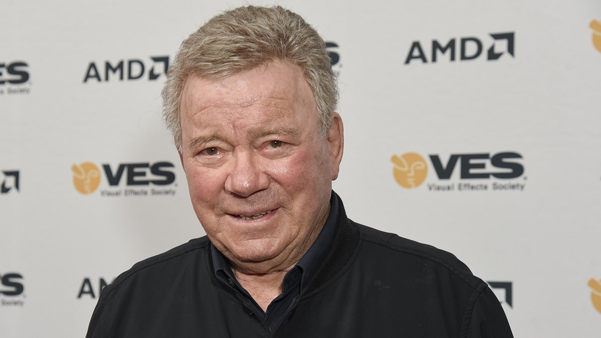 William Shatner in a black button down smiles on the carpet