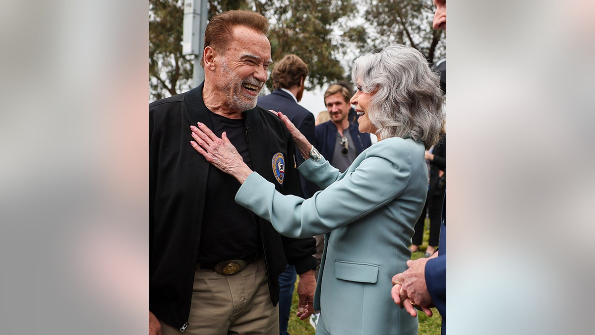Arnold Schwarzengger sorri em seu terno preto e calça cáqui bege enquanto é recebido por Jane Fonda em um terno azul