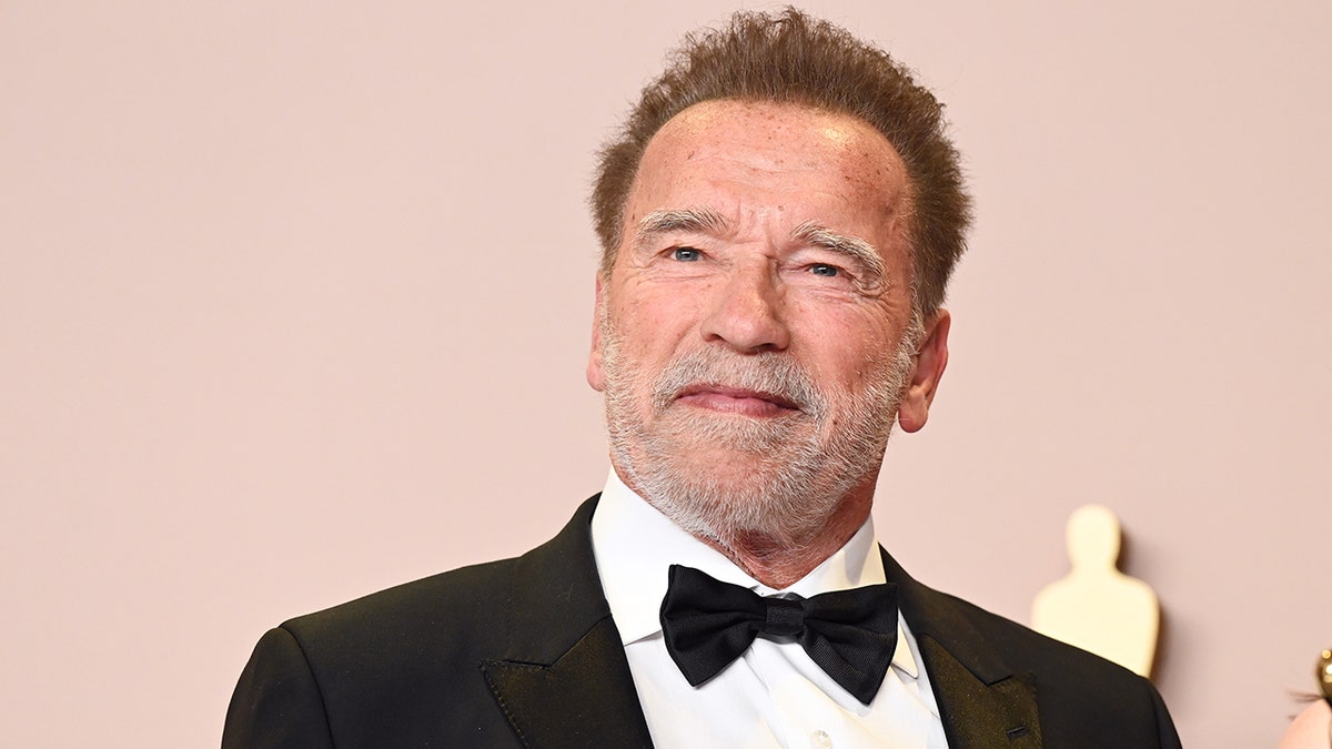 Arnold Schwarzenegger em um smoking preto sorri suavemente no tapete do Oscar