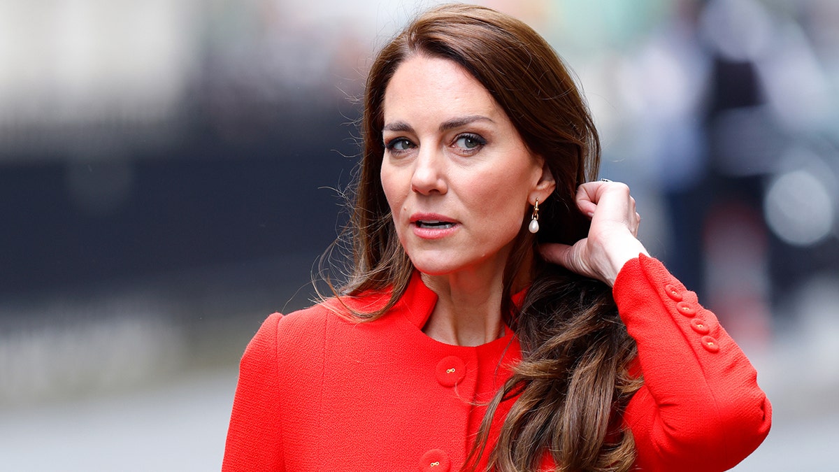Kate Middleton in a bright red orange suit pulls her hair back as she looks away from the camera
