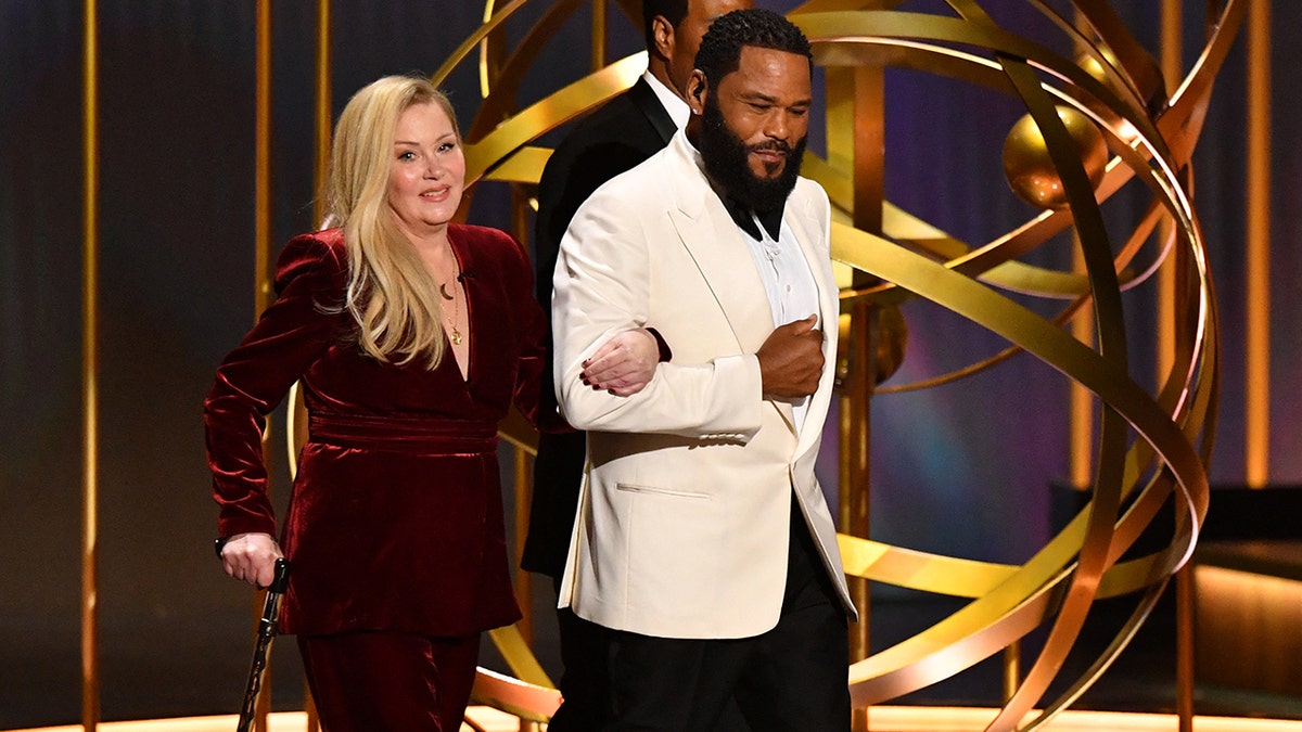 Christina Applegate in a red velvet dress and using a cane is led on stage by Anthony Anderson in a off-white tuxedo on Emmy's stage