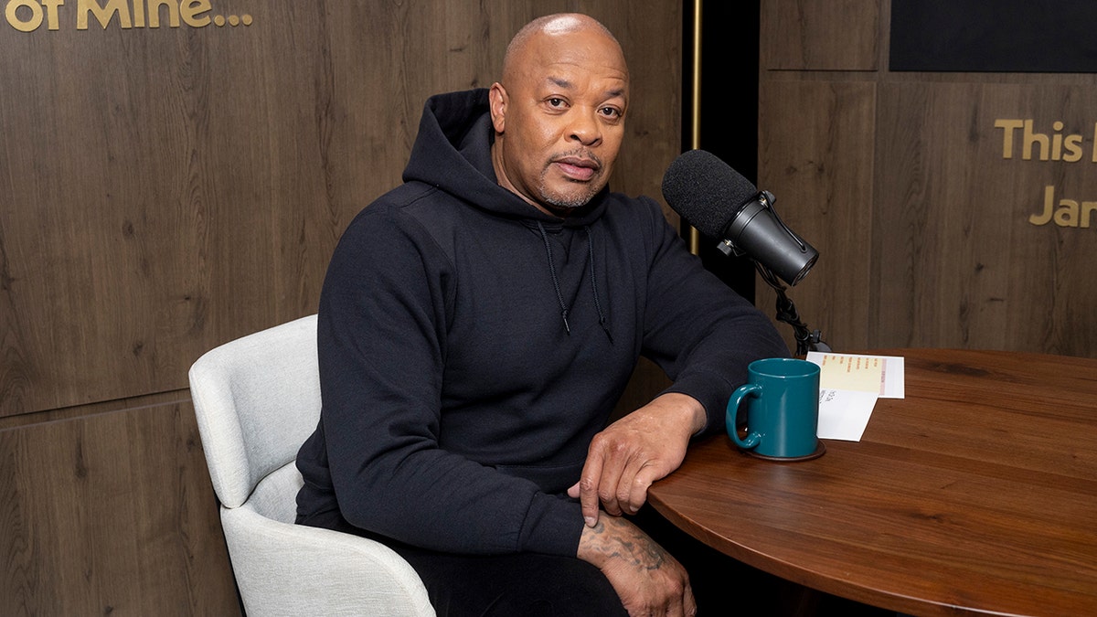 Dr. Dre in a black sweatshirt sits in a white chair at a large brown table and smiles for a photo