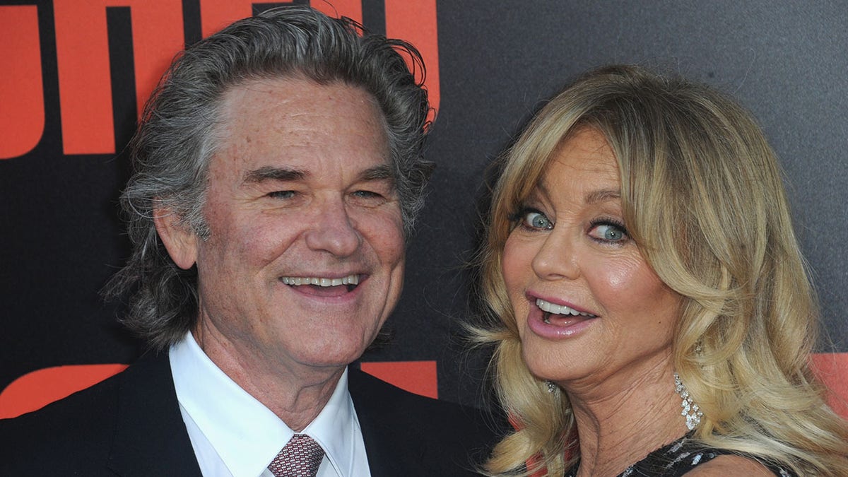 Kurt Russell de terno preto sorri ao lado de Goldie Hawn, que faz uma cara de surpresa/boba no tapete