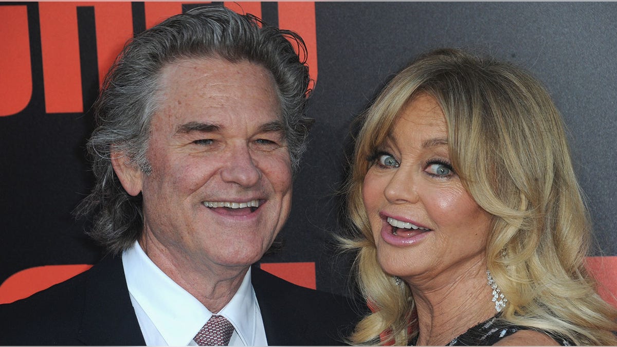 Kurt Russell in a black suit smiles next to Goldie Hawn who makes a surprised/silly face on the carpet