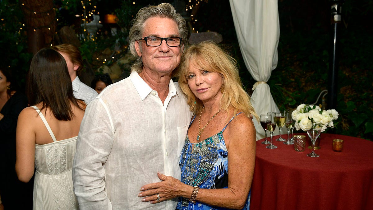 Kurt Russell in a white shirt and Goldie Hawn in a blue printed dress pose for a photo