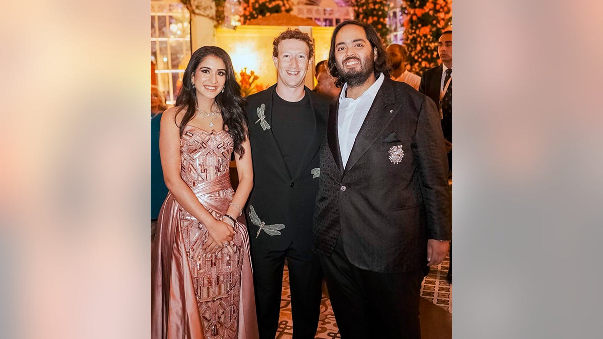 Radhika in a rose gold pink sparkly dress stands next to Mark Zuckerberg in a black blazer with dragonflies on it next to Anant Ambani in a dark tuxedo jacket