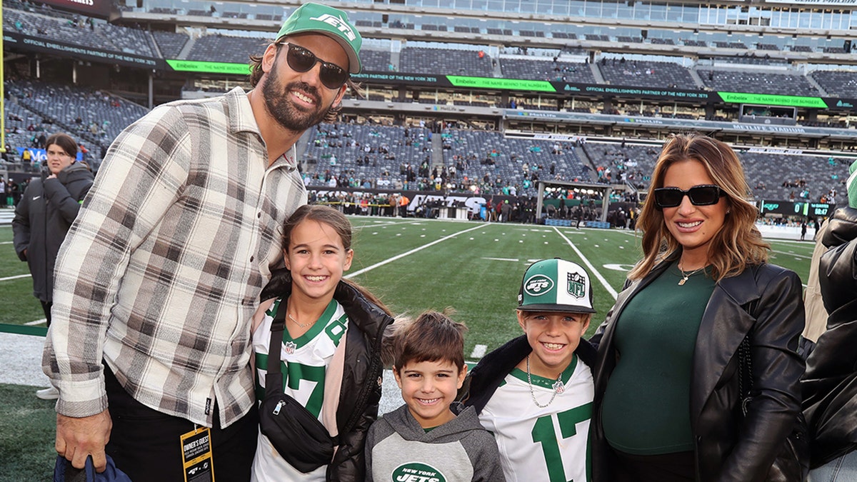 Eric Decker in a flannel shirts and green Jets hat smiles with his children Vivianne, Forrest and Eric and pregant wife Jessie James Decker in green