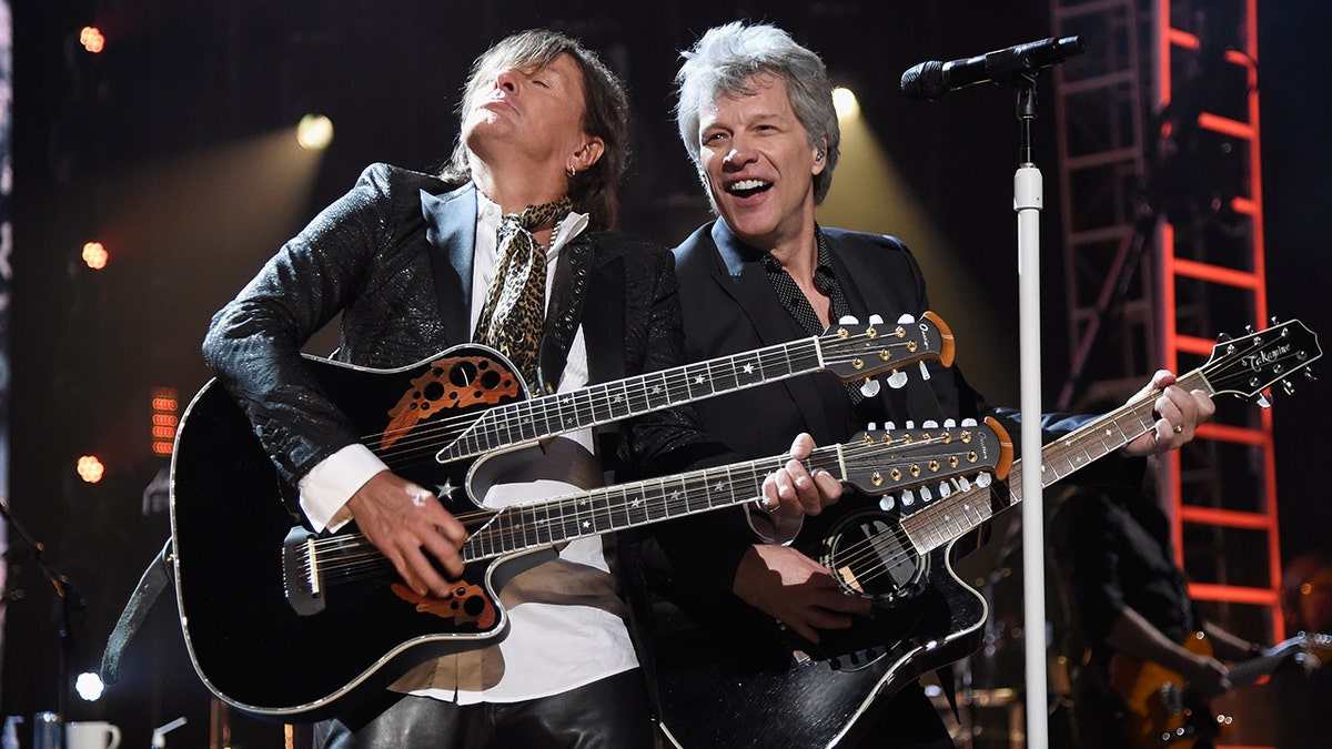 Richie Sambora joga a cabeça para trás e toca guitarra enquanto Jon Bon Jovi ri e observa também tocando