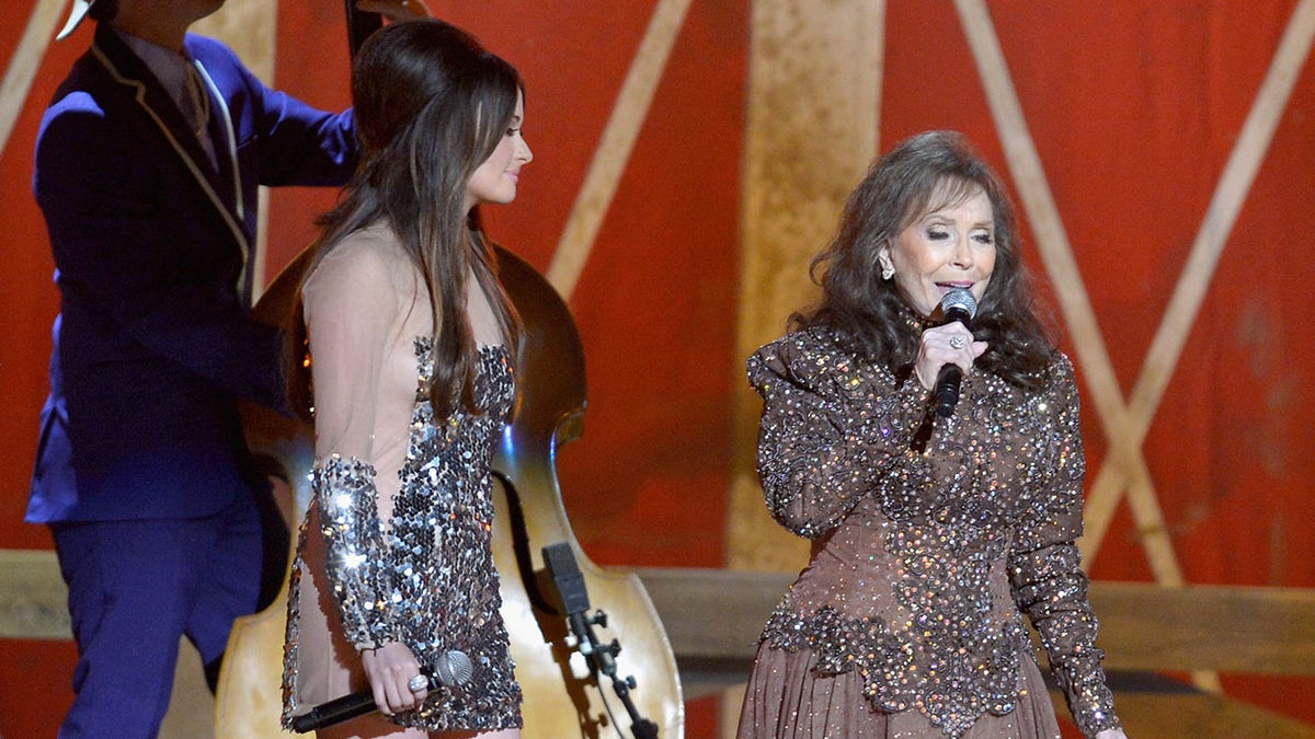 Kacey Musgraves wearing a sheer and sparkly dress on stage next to Loretta Lynn who sings into the microphone