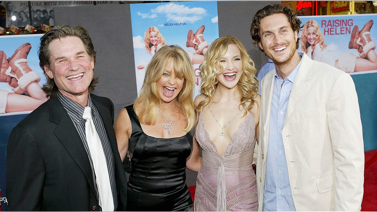 Kurt Russell in a black suit and black and white striped shirt laughs with Goldie Hawn ina. black dress, Kate Hudson in a light pink lacy dress and Oliver Hudson in a cream jacket on the carpet