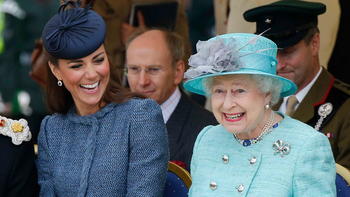 Kate Middleton con traje azul y sombrero a juego se ríe sentada junto a la Reina Isabel con traje turquesa pálido y sombrero a juego