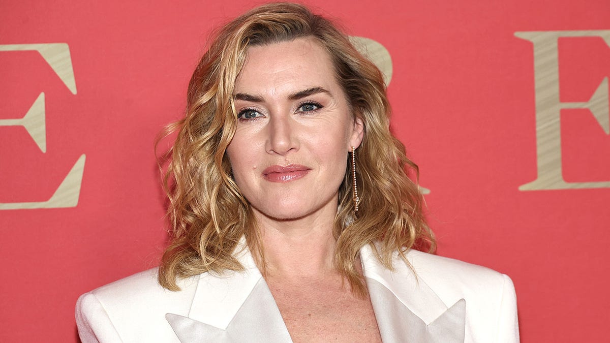 Kate Winslet in a white blazer looks to her left and soft smiles on the carpet
