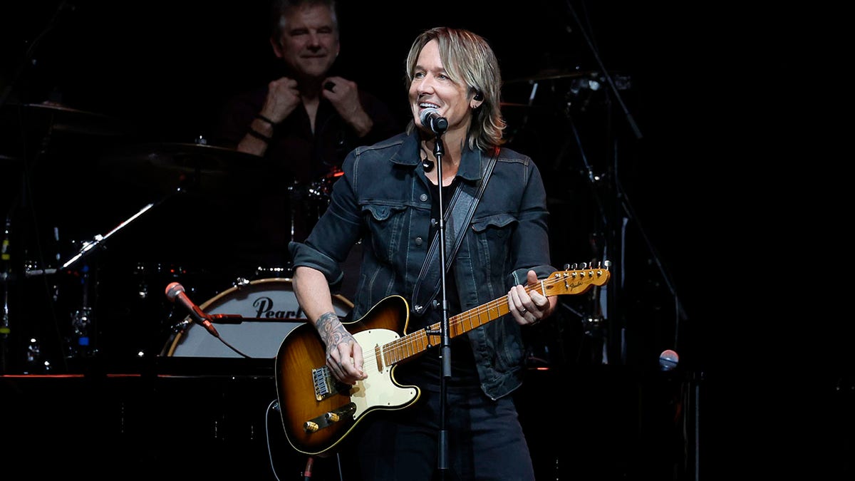 Keith Urban in a jean jacket plays the guitar on stage and looks to his right