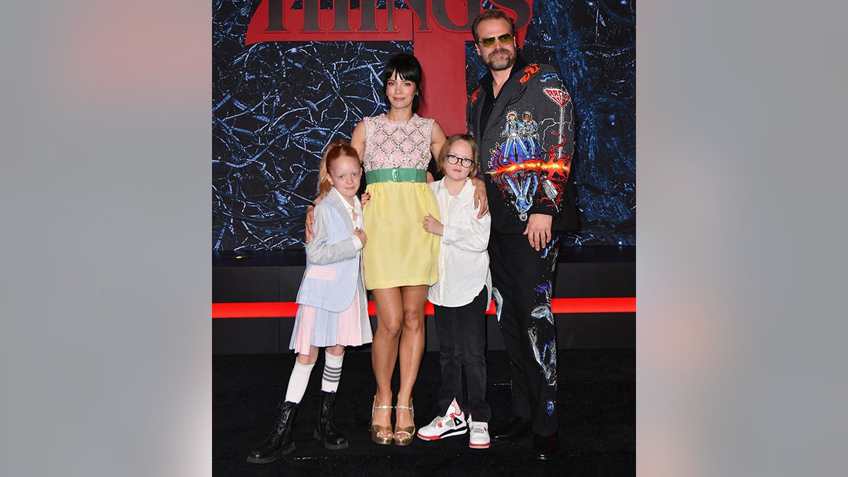 Lily Allen in a multicolored dress with a yellow skirt and green belt is held by her two daughters on the carpet with husband David Barbour