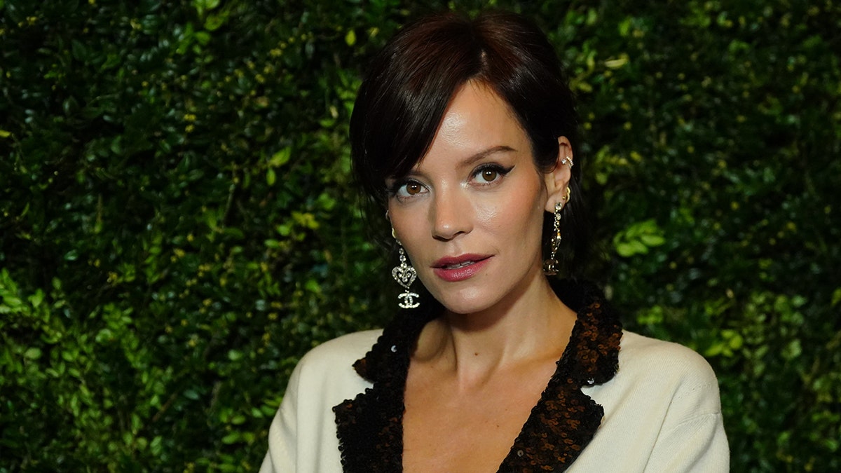 Lily Allen looks up and away from the camera in a white blazer with black trim and dangly Chanel earrings