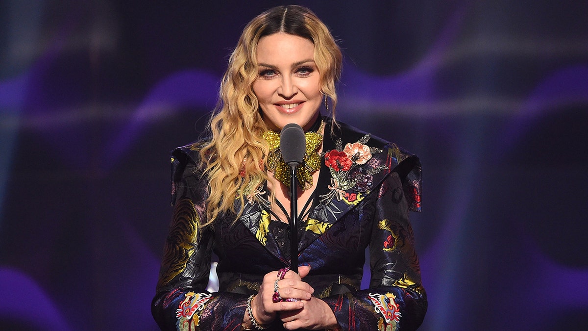 Madonna holds a microphone on stage while wearing colorful floral blazer