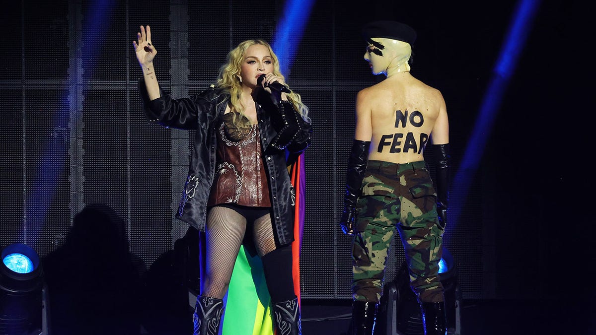 Madonna in a lacy black leotard and tights and dark black jacket singing on stage standing next to a dancer that has the words 'No Fear' written on their back