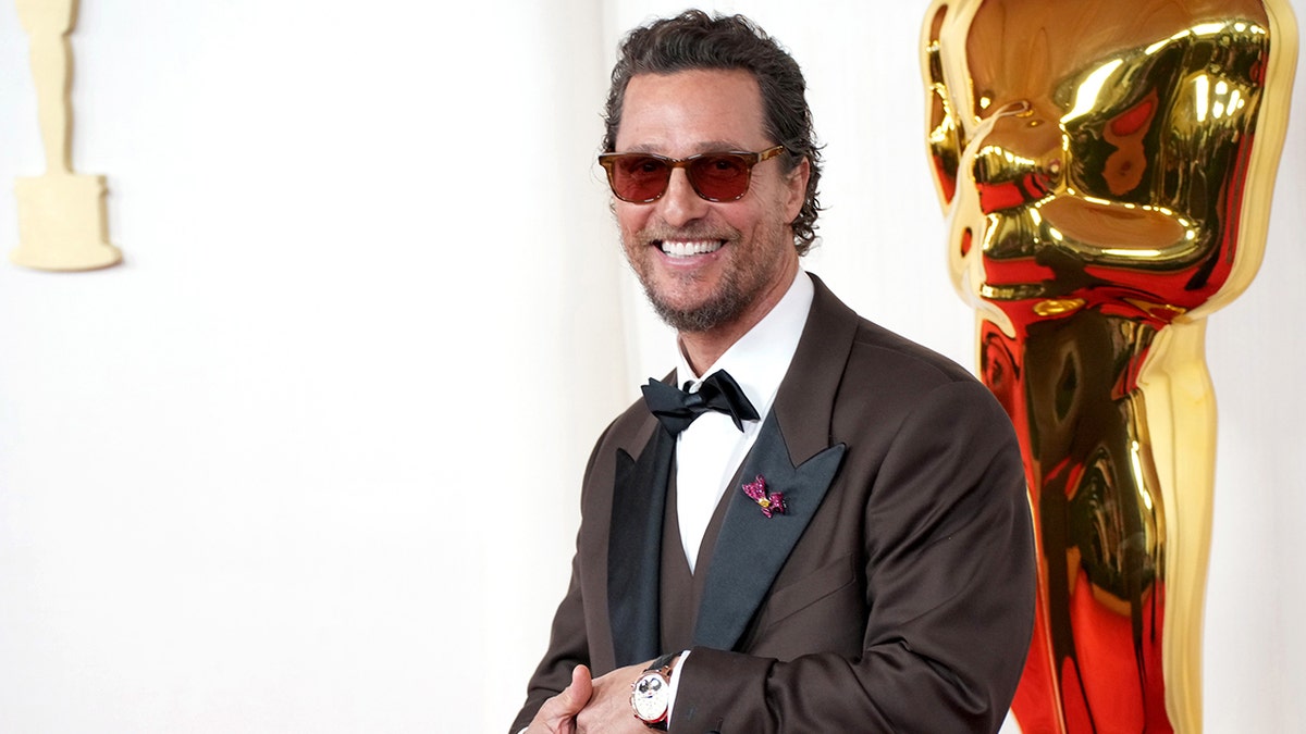 Matthew McConaughey in a brown tuxedo smiles with his glasses on at the Oscars