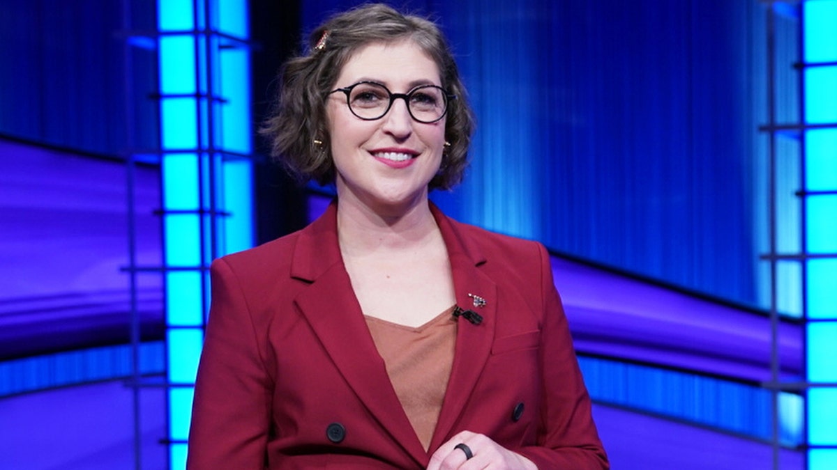 Mayim Bialik in a red pantsuit hosting Jeopardy!