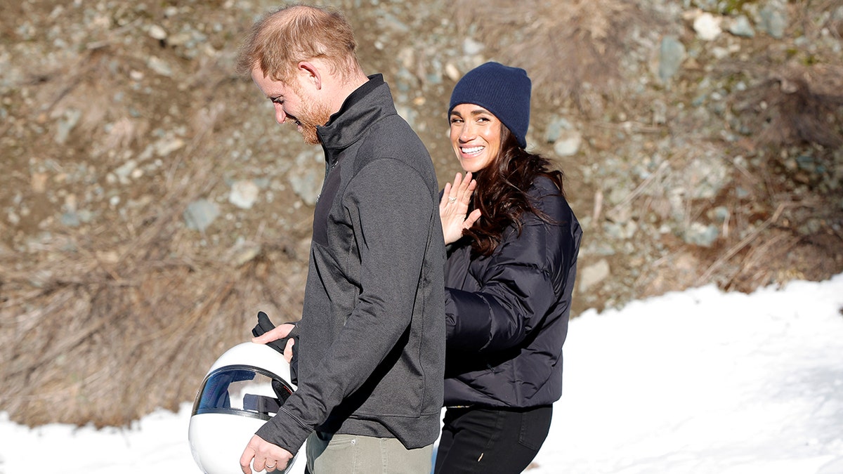 Prince Harry walks with Meghan Markle in a blue beanie who turns to the cameras and waves in British Columbia