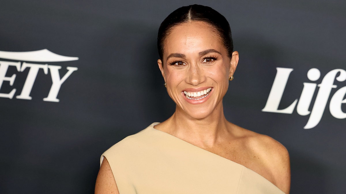 Meghan Markle tersenyum di atas karpet dengan gaun one-shoulder berwarna krem