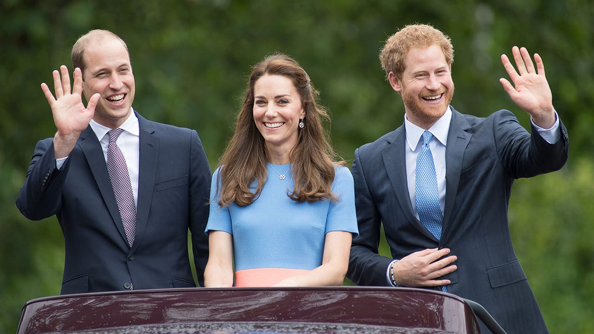 Prince William waves in a navy blue suit next to Kate Middleton in a light blue dress next to Prince Harry in a navy suit, sticking out the roof of a vehicle
