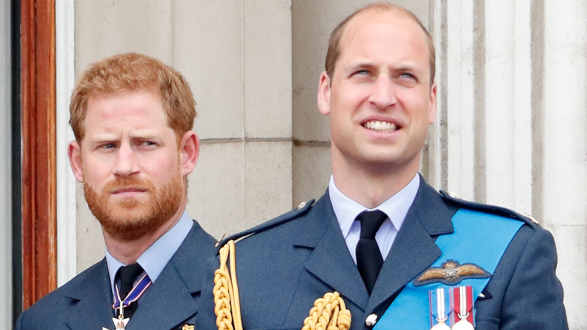 A tense Prince Harry stands behind his brother Prince William, smiling, on the royal balcony