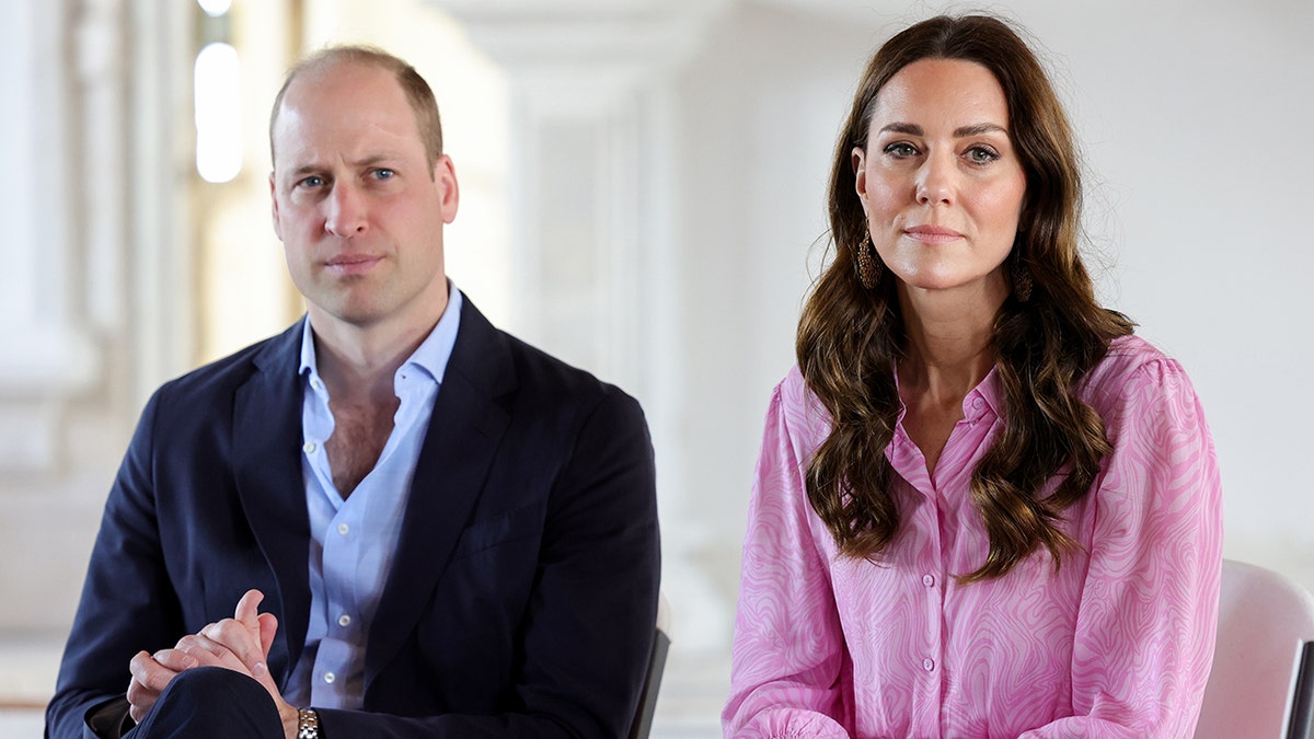 Prince William in a black suit over a light blue shirt sits and looks serious next to Kate Middleton in a pink outfit, also looking stoic