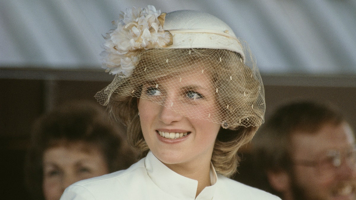 Princess Diana in an off white outfit and matching hat with a slight vail over it with pearls smiles as she looks away from the camera