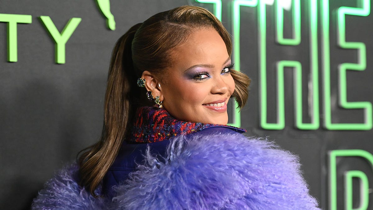 Rihanna looks behind her on the carpet wearing a shaggy purple jacket and smiles