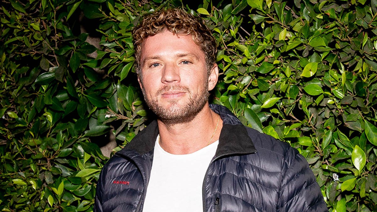 Pyan Phillippe soft smiles in a dark puffer jacket and white shirt in front of ivy backdrop