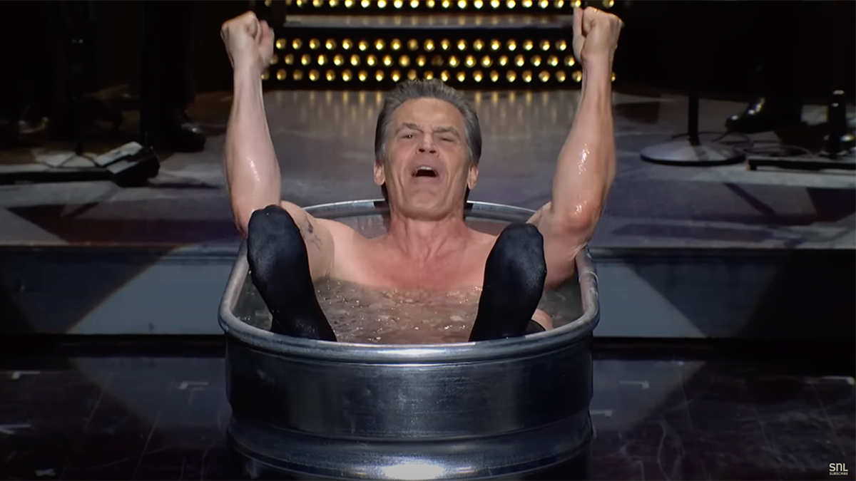 Josh Brolin in a tub doing a cold plunge on the set of "SNL" with his arms raised up and his feet sticking out