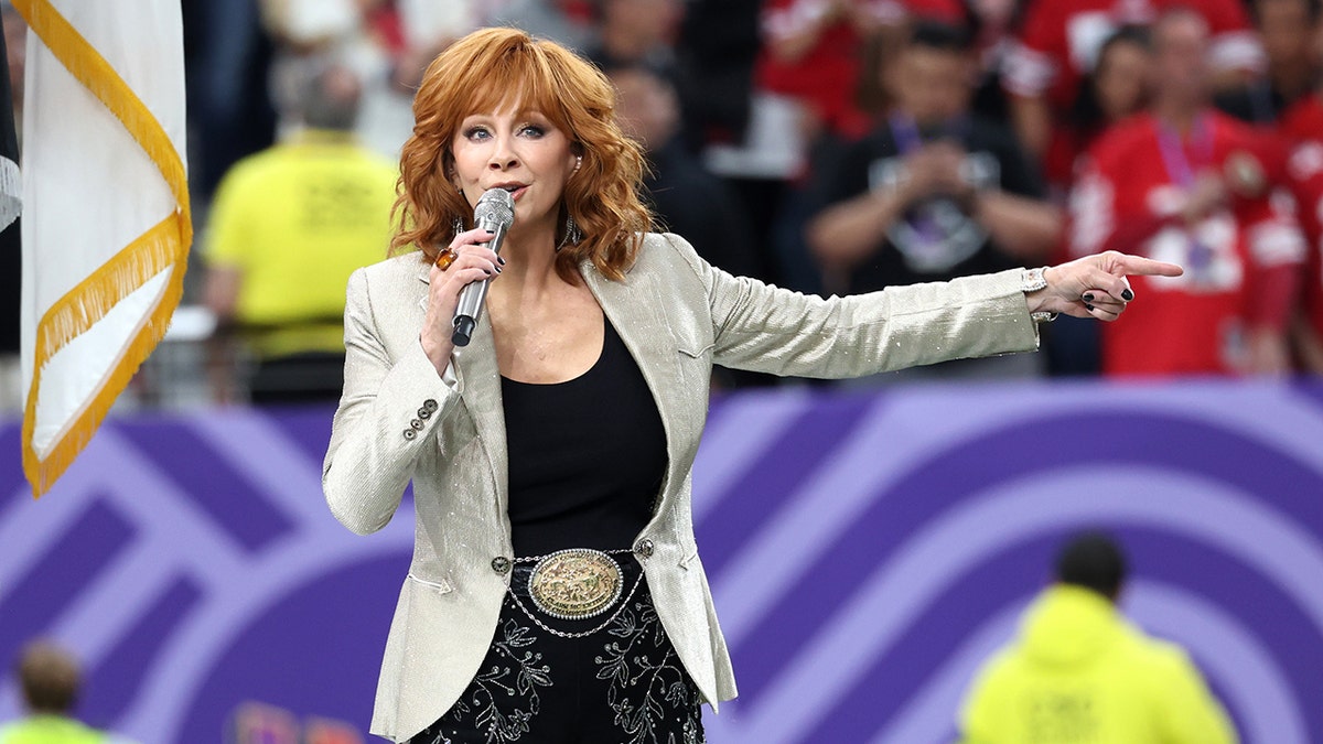 Reba McEntire stretches out her left arm and holds her microphone to her mouth with her right while peforming the Star Spangled Banner at the Super Bowl, wearing a light sand colored jacket and black shirt with a massive belt buckle