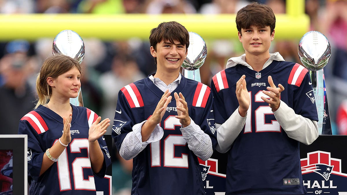 Vivian and Benjamin Brady and Jack Moynihan clap at a ceremony for their father Tom Brady at Gillette Stadium, all wearing their dad's jersey