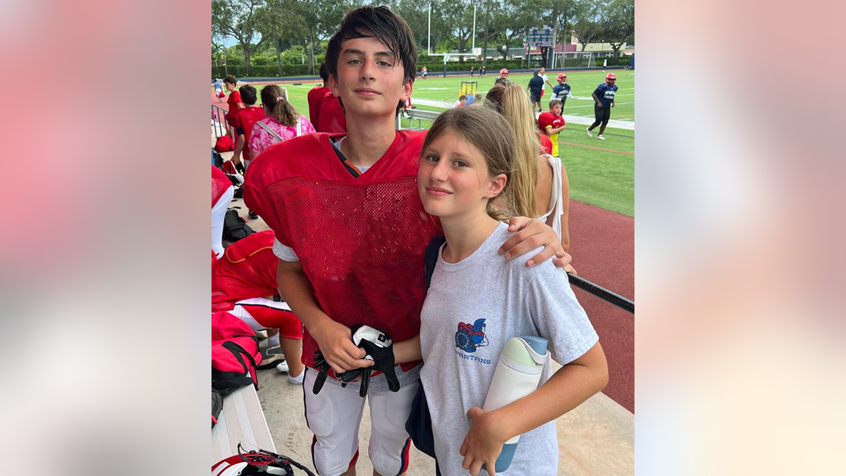 Benjamin Brady in a red shirt at a football game/practice stands tall next to his sister Viivan in a white t-shirt 