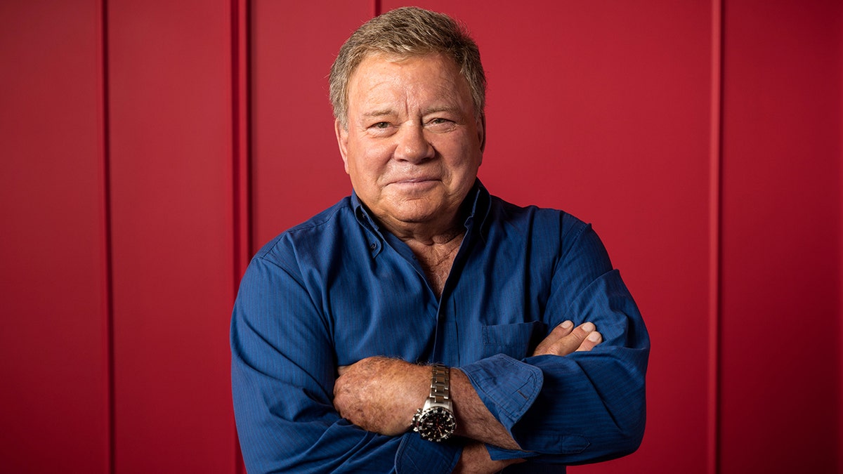 William Shatner in a blue shirt crosses his arms over his chest and soft smiles
