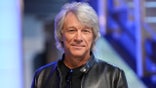 Jon Bon Jovi soft smiles and looks directly into the camera wearing a leather jacket