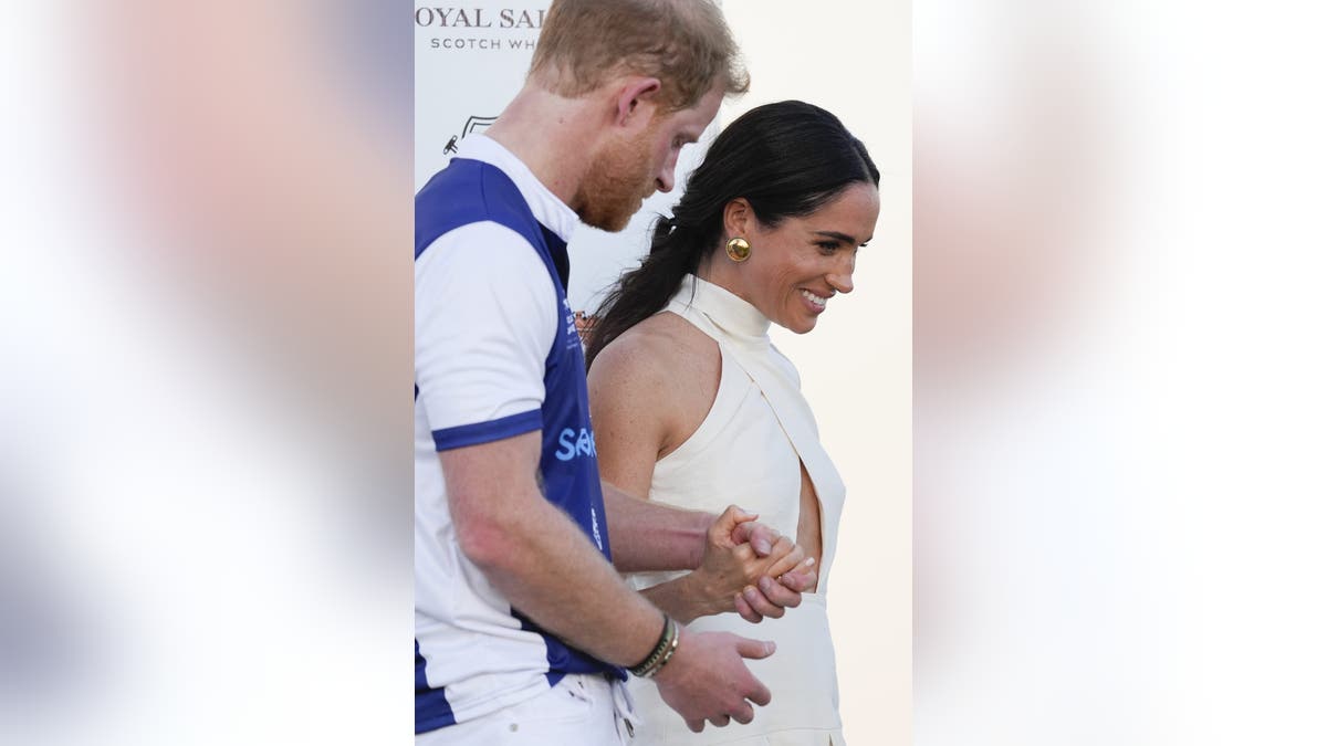 Prince Harry in a polo suit holding Meghan Markle's hand as she wears a cut-out white dress.