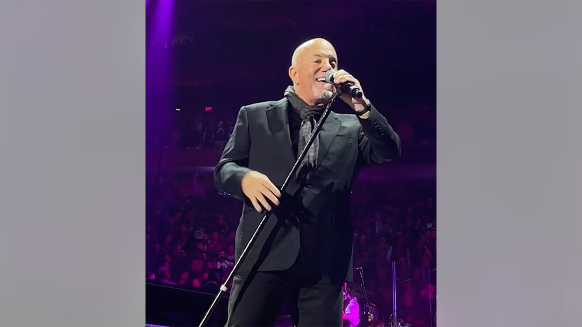 Billy Joel sostiene su micrófono en el aire en el Madison Square Garden mientras interpreta "Uptown Girl" y sonríe