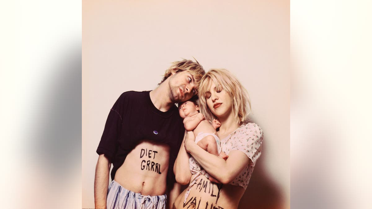 Kurt Cobain e Courtney Love carregando seu bebê em frente a uma parede bege.