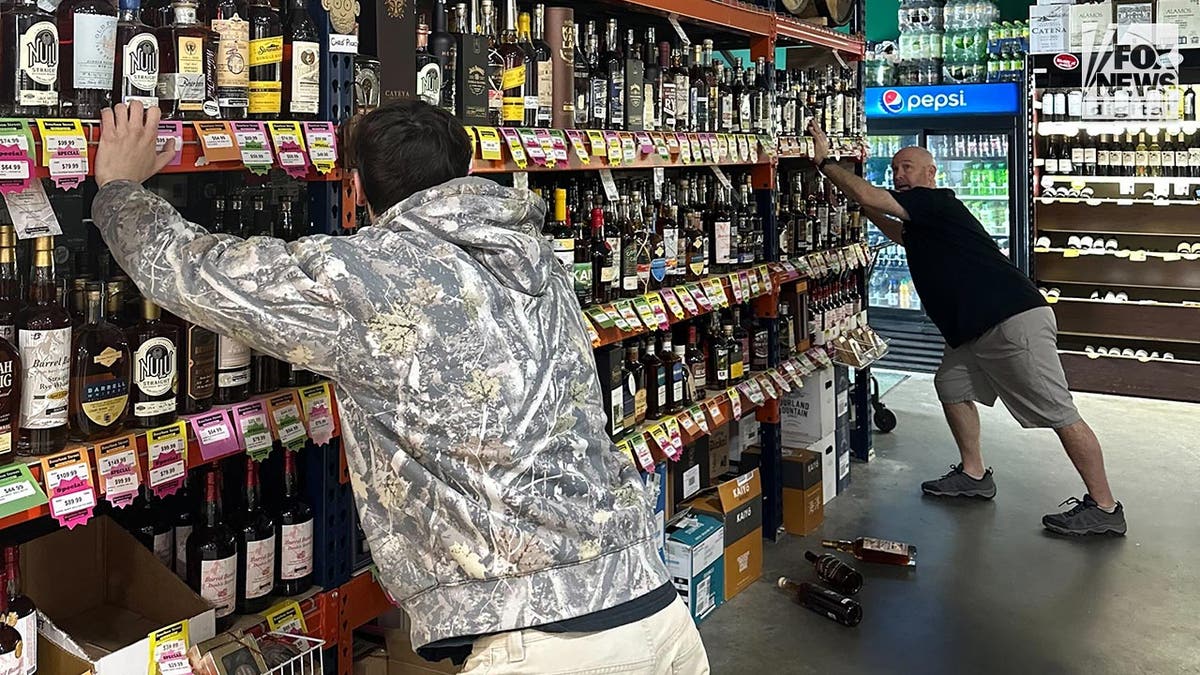 Two men prevent a shelf containing alcohol bottles from shaking