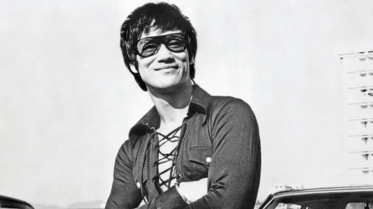 Um close de Bruce Lee sorrindo e usando óculos escuros